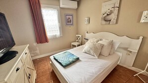 Un dormitorio con cama, televisor, silla, mesita y ventana con cortinas.