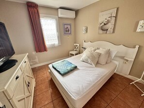 Ein Schlafzimmer mit Bett, Fernseher, Sessel, kleinem Tisch und einem Fenster mit Vorhängen.