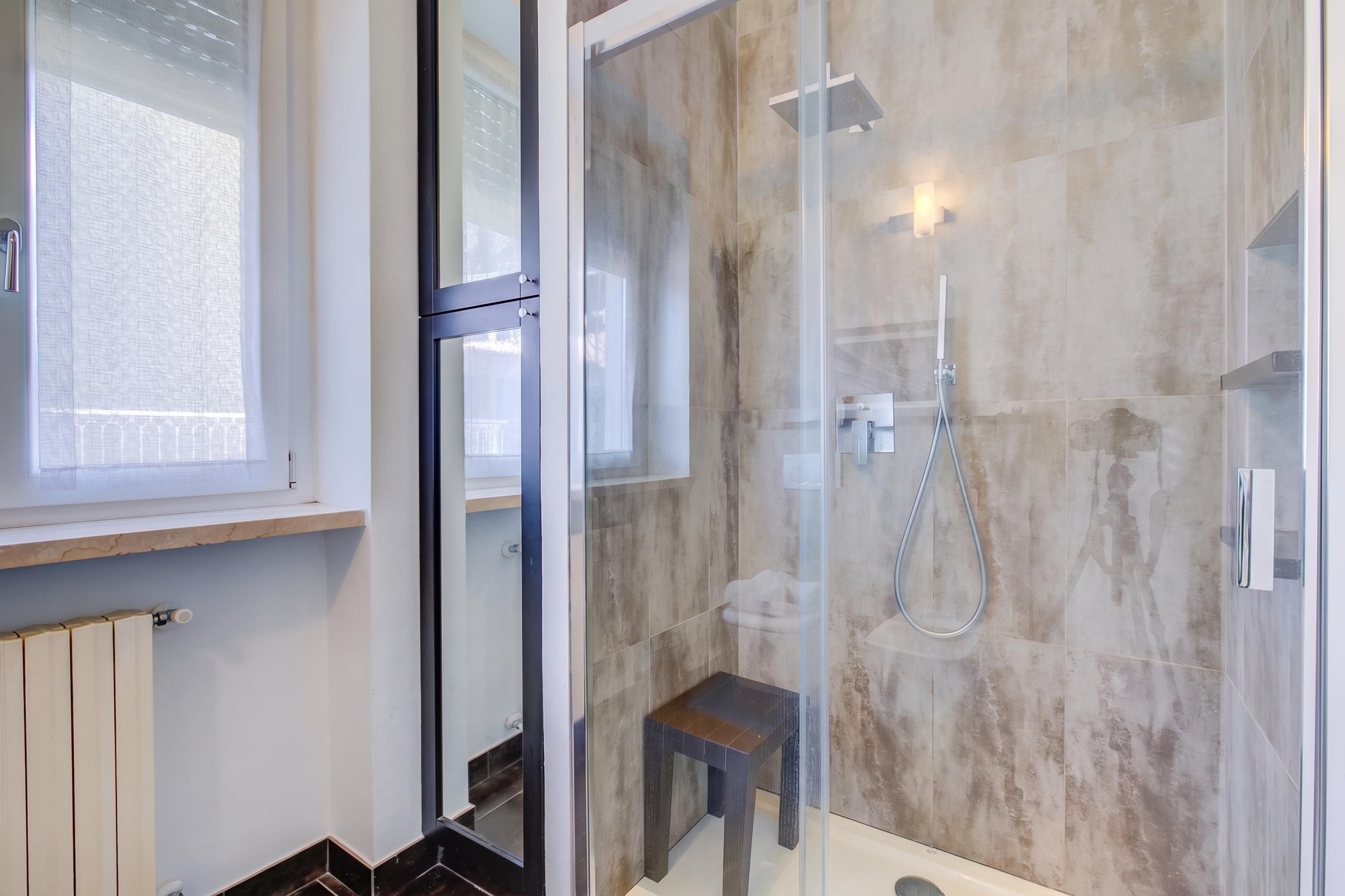 A modern bathroom with a glass shower enclosure, a wooden bench, and a visible window.