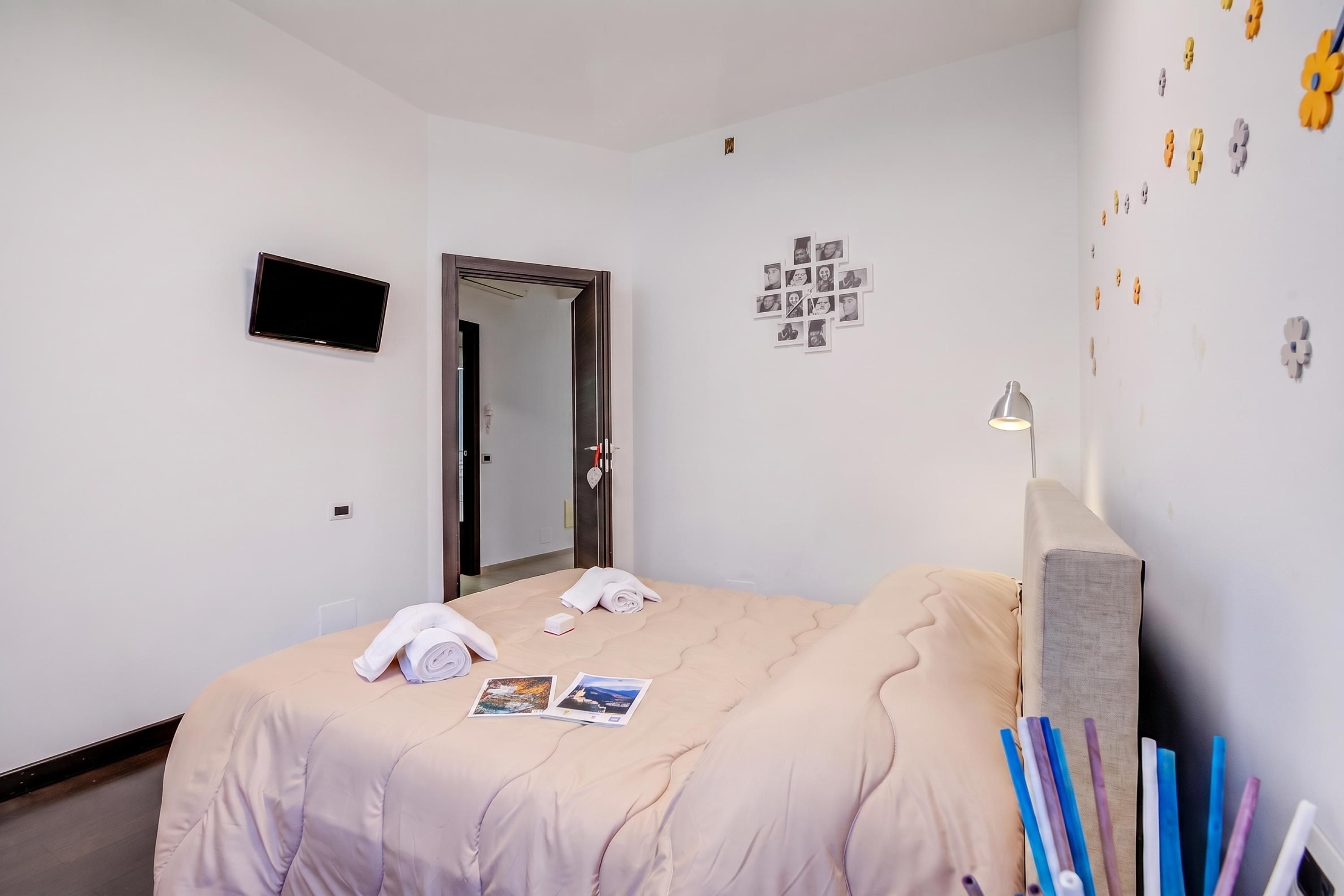Habitación de hotel con cama, televisión de pantalla plana, espejo, reloj de pared y mesita de noche con lámpara.