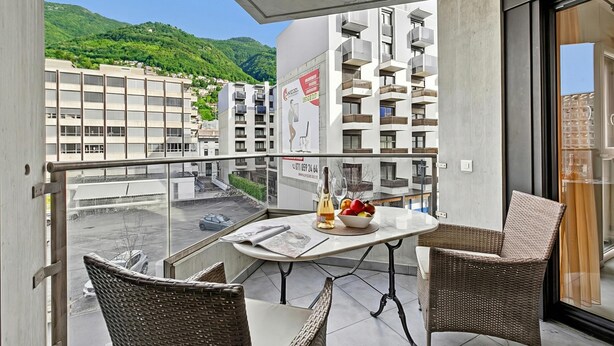 A balcony with wicker chairs, a table with a newspaper, a bottle, and a bowl of fruit. - Locarno Centro Apartment 2BR With Private Garage (Locarno)