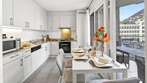 A modern kitchen with white cabinets, a dining area with a table set for two, and a view of the city through large windows. - Locarno Centro Apartment 2BR With Private Garage (Locarno)