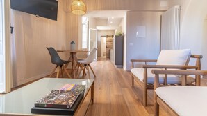 Una sala de estar moderna con techo de madera, una mesa de centro de vidrio con revistas, un televisor montado en la pared y un comedor con sillas y una mesa.