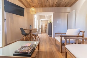 A modern living room with a wooden ceiling, a glass coffee table with magazines, a TV mounted on the wall, and a dining area with chairs and a table. - Hortus apartment in the historic centre (Orta San Giulio)