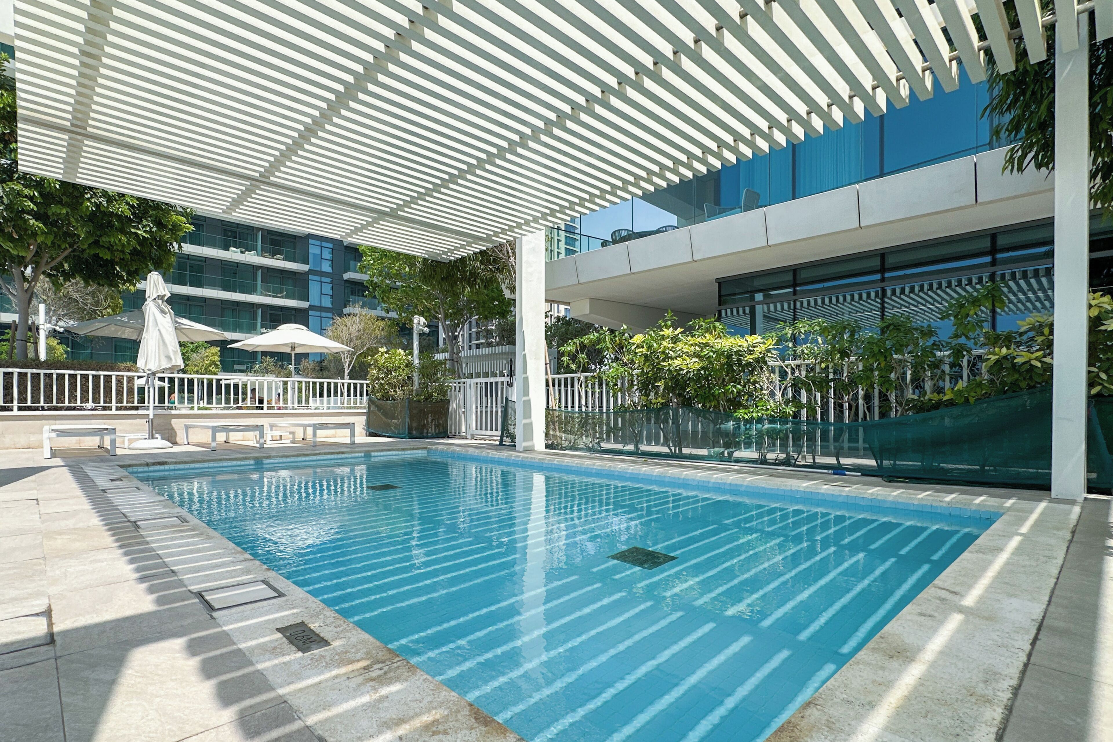 A swimming pool with a covered area, lounge chairs, and a view of a modern building.