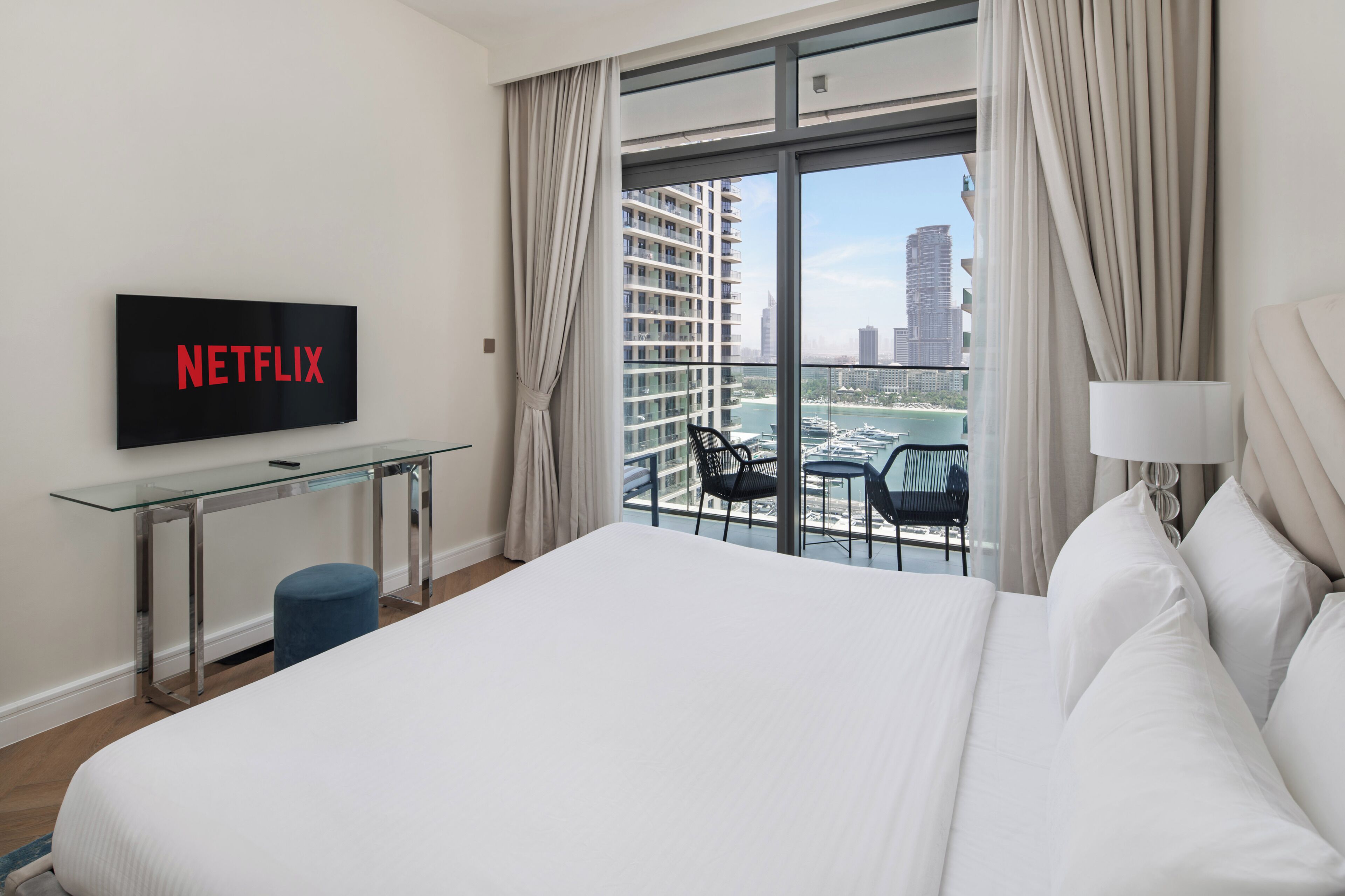 Habitación de hotel con cama, televisor mostrando 'NETFLIX', una mesita de vidrio, una silla y vistas de un paisaje urbano a través de amplias ventanas.