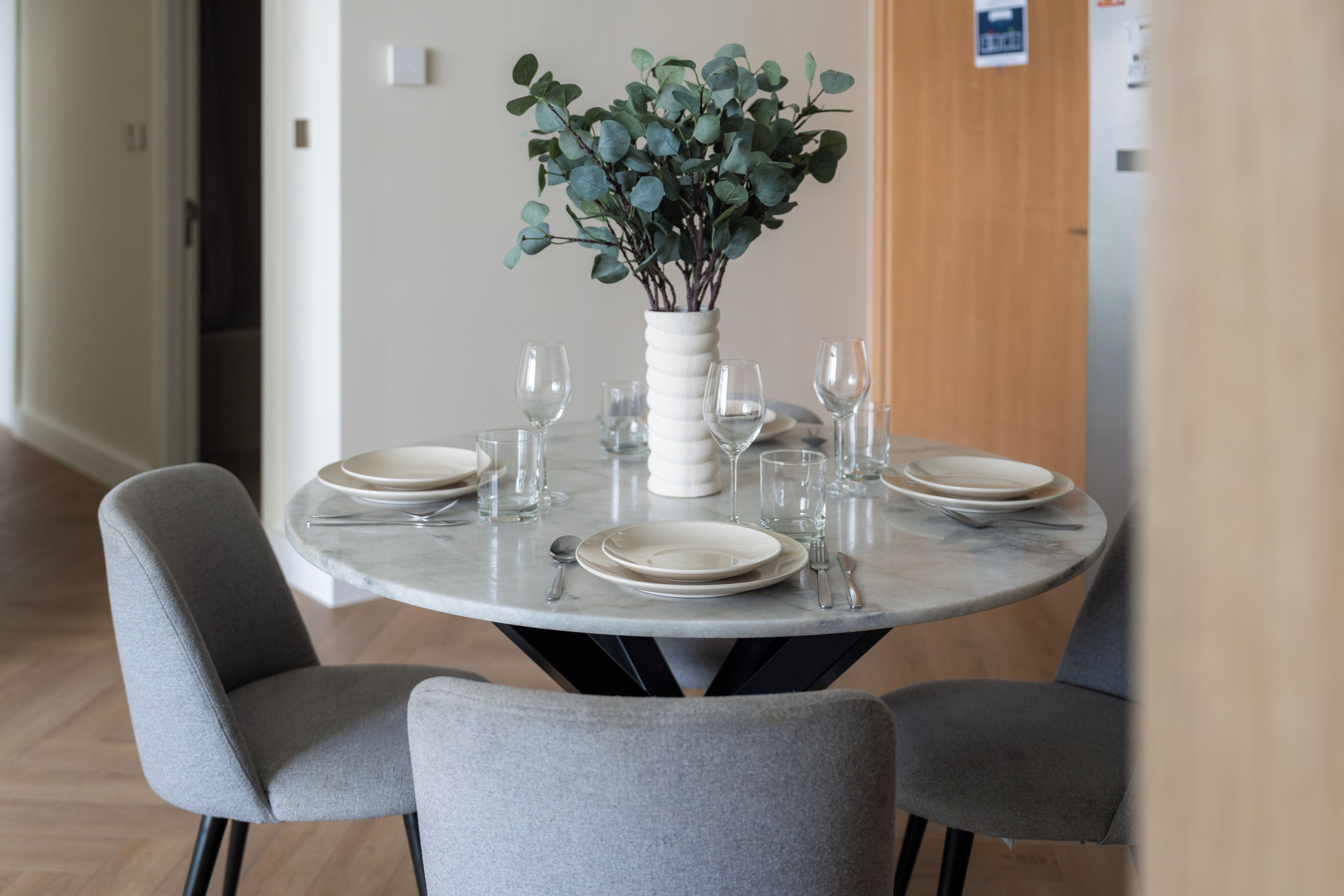 A round dining table with four chairs, a vase with green foliage, and empty dinnerware.