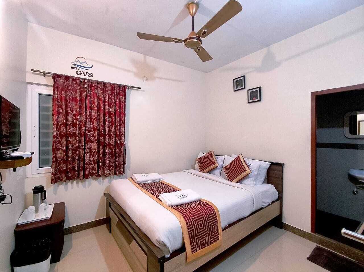 A hotel room with a bed, a ceiling fan, a window with curtains, a bedside table with a coffee maker, and a TV.