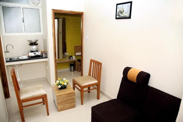A compact living space with a kitchenette, a wooden chair, a sofa, and a small table with a flower arrangement. - Apollo Court (Chennai)