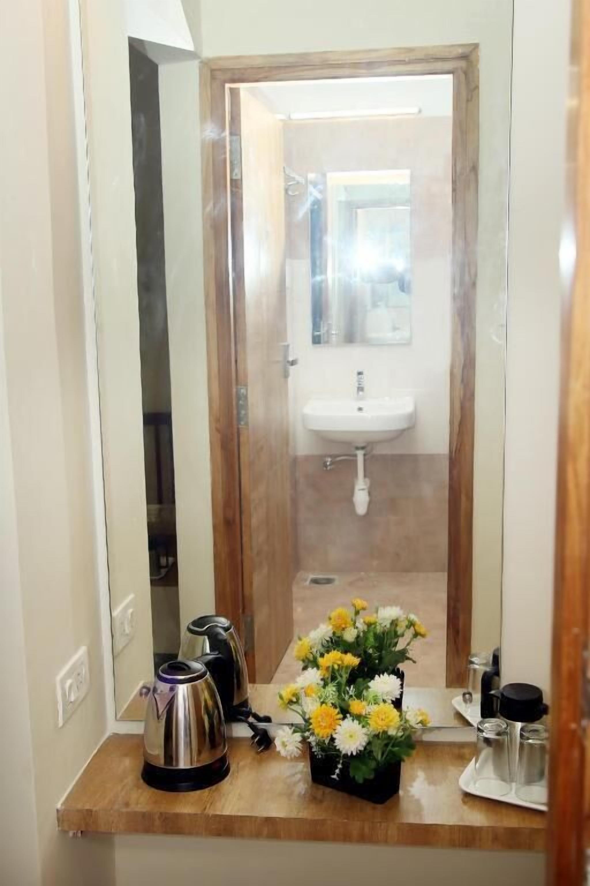 Baño con puerta de madera, un lavamanos y un espejo. Hay una cafetera, un hervidor y un arreglo floral en la encimera.