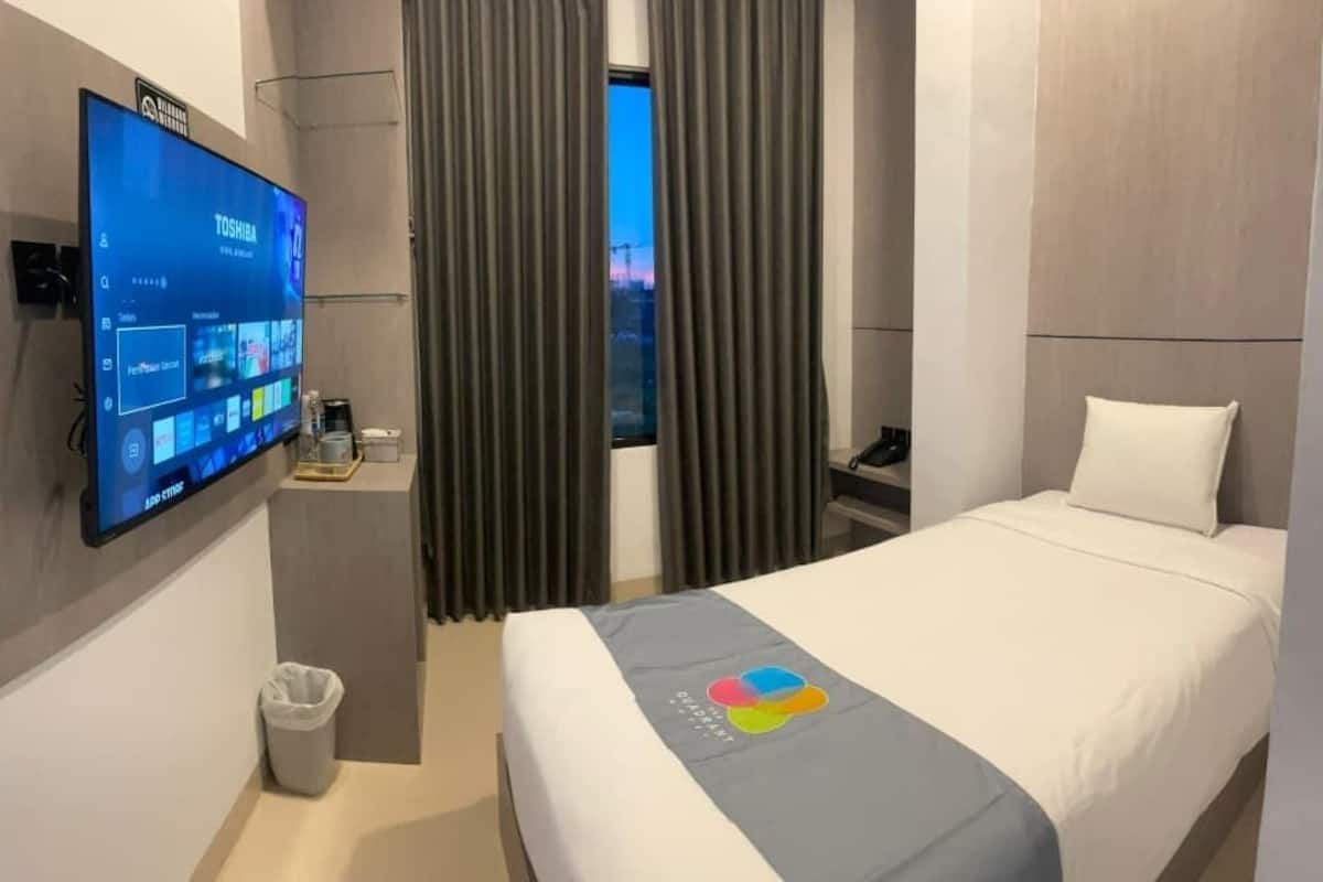 Una habitación de hotel con cama, televisión montada en la pared, ventana con cortinas y un cubo de basura pequeño.
