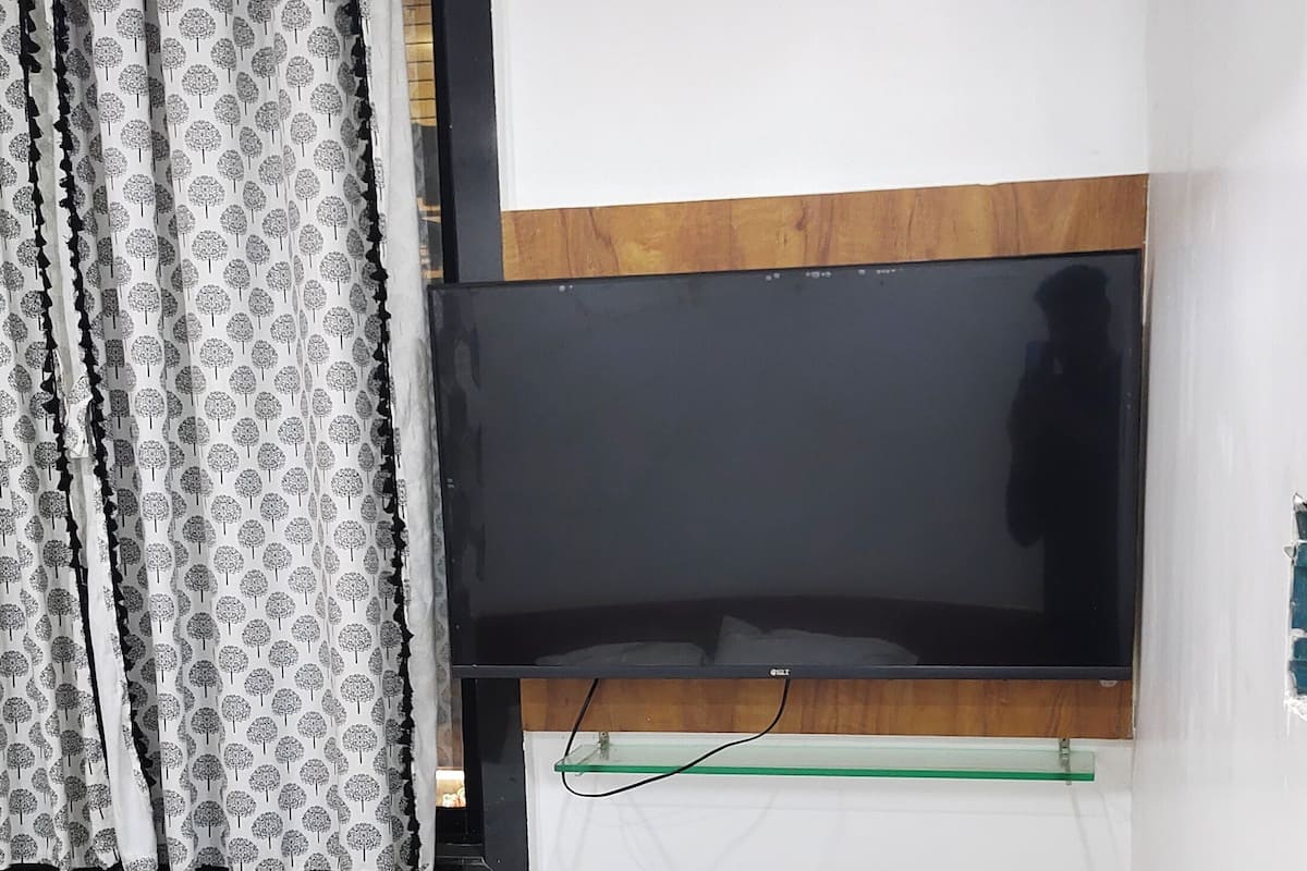 A room with a bed, a wooden side table, a mounted TV, and patterned curtains.