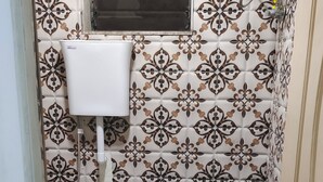 Un baño pequeño con azulejos estampados, un inodoro, una ventana y un cubo azul.
