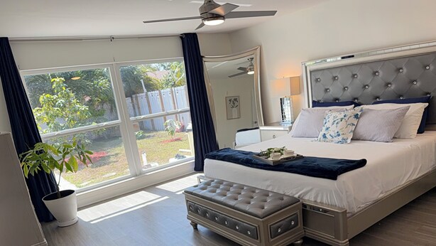 A bedroom with a large bed, a ceiling fan, a potted plant, and a view of the outdoors.