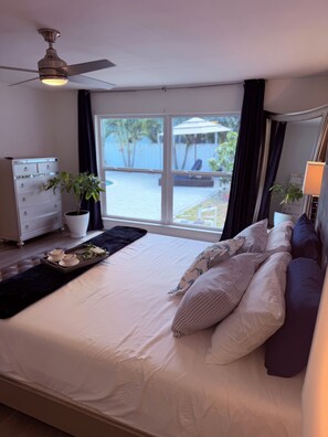 Un dormitorio con cama, ventilador de techo, ventana con vista a una piscina, cómoda y una planta.