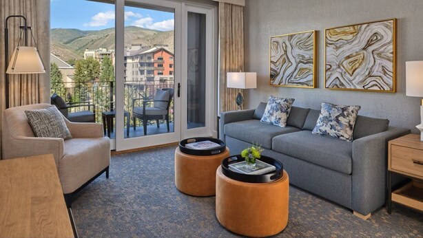 Ein Hotelzimmer mit grauem Sofa, zwei runden Beistelltischen, einem Holztisch und einem Balkon mit Blick auf Gebäude und Hügel. An der Wand hängt ein abstraktes Kunstwerk.