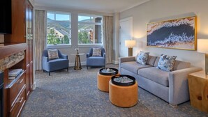 Ein Hotelzimmer mit einer Couch, zwei Sesseln, einem Couchtisch, einem Fernseher, einem Gemälde und Blick auf Gebäude durch das Fenster.
