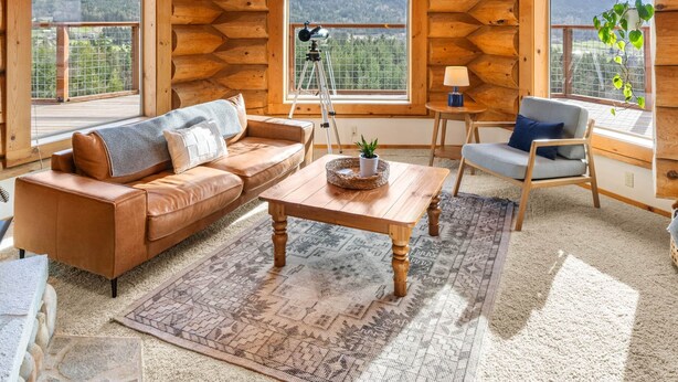 Een blokhutinterieur met een hoge plafond, houten wanden, een plafondventilator, een bank, een tafel en grote ramen met uitzicht op de bergen.