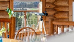 Het interieur van een blokhut met een telescoop op een statief, houten wanden, een raam met uitzicht op de natuur, en een eettafel met stoelen.