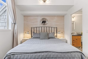 Une chambre à coucher comprenant un lit, deux tables de chevet, un luminaire fixé au mur, une tête de lit en bois et une fenêtre avec des rideaux.