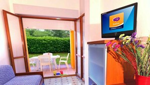 Ein kompakter Raum mit Bett, einer kleinen Küchenzeile, einem Fernseher, der 'Carina LIVENZARESORTS' anzeigt, und einer Glastür, die zu einem Außenbereich mit Tisch und Stühlen führt.