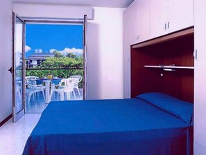 A room with a bed, a balcony with a table and chairs, and a view of buildings and greenery. - Holiday apartment with air conditioning, balcony and pool (Bibione)