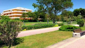 A multi-story hotel with a flat roof and balconies, surrounded by well-maintained greenery and a paved walkway. - Holiday flat with air conditioning and balcony (Bibione)