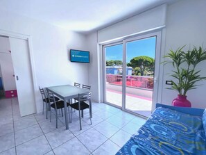 Ein Zimmer mit Esstisch und Stühlen, einem an der Wand montierten Fernseher, einer großen Schiebeglasdoor zum Außenbereich und einem Blumentopf.