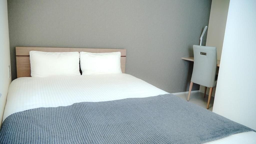 A neatly made bed with white linens and a gray throw blanket, a wooden headboard, and a small desk with a chair to the right.