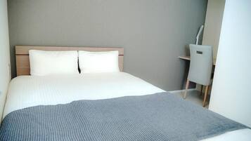 A neatly made bed with white linens and a gray throw blanket, a wooden headboard, and a small desk with a chair to the right.