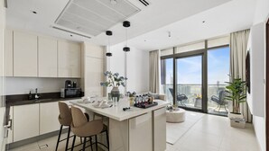 A modern kitchen with a central island, a dining area with chairs, a large window with a city view, and a balcony with a chair. - Deluxe Marina Gate Suite w/ Sea & Ain Dubai View (Dubai)