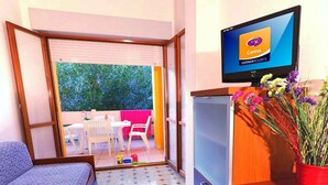 Quarto compacto com cama, pequeno espaço de cozinha, TV exibindo o logotipo Carina e varanda com mesa e cadeiras.