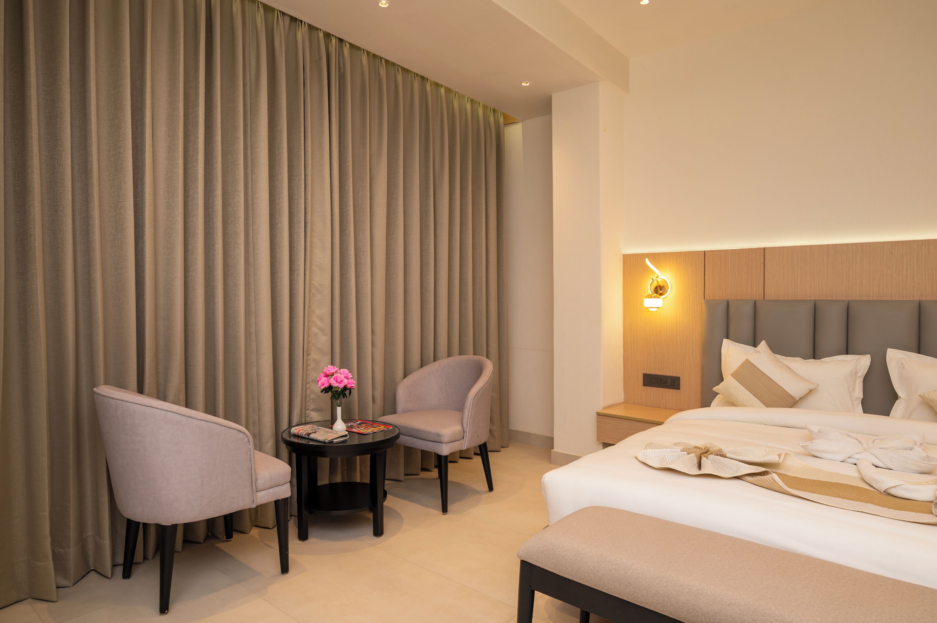 Habitación de hotel con una cama, dos sillones, una mesita con un jarrón y flores, y una mesita de noche con una lámpara.