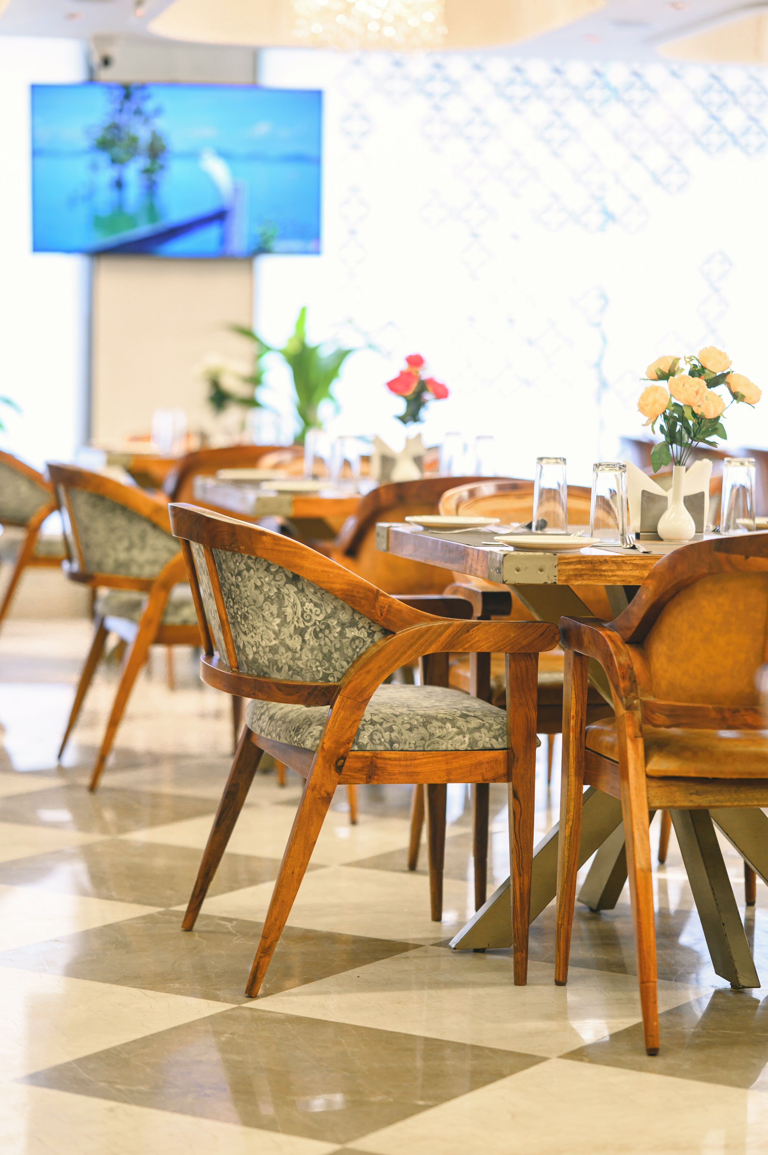 A dining area with wooden tables and chairs, a flat-screen TV mounted on the wall, and a patterned wall in the background.