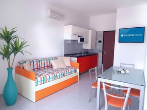 A compact living space with a kitchenette, a sofa, a dining table with chairs, and a TV mounted on the wall. - Holiday apartment with air conditioning, balcony and pool (Bibione)