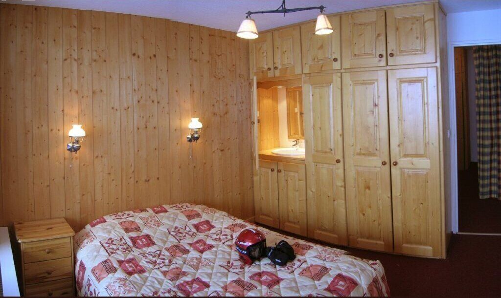 Habitación con paneles de madera, una cama con colcha estampada, dos lámparas fijadas a la pared y un armario de madera con espejo.