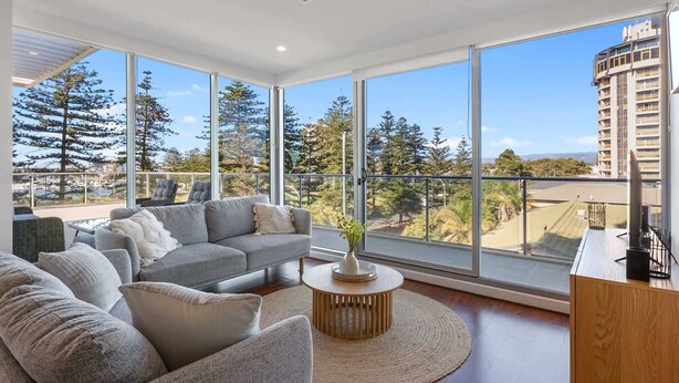 A spacious living room with a grey sectional sofa, a round wooden coffee table, and a view of a cityscape through large windows. - Stylish 2-Bed with Terrace, 3-min Walk to Beach (Glenelg)