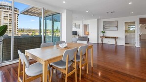 A spacious living area with a wooden dining table, grey upholstered chairs, a sofa, and a large window offering a view of a cityscape. - Stylish 2-Bed with Terrace, 3-min Walk to Beach (Glenelg)