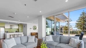 A modern living room with two grey sofas, a coffee table with vases, a kitchen in the background, and a large window with a view of buildings and trees. - Stylish 2-Bed with Terrace, 3-min Walk to Beach (Glenelg)