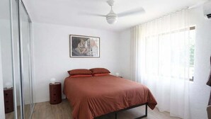 Um quarto com cama, ventoinha no teto, mesa de cabeceira, espelho e janela com cortinas.