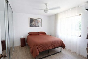 Ein Schlafzimmer mit einem Bett, einem Deckenventilator, einem Nachttisch, einem Spiegel und einem Fenster mit Vorhängen.