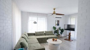 Uma sala de estar moderna com um sofá seccional, uma mesa de centro redonda, um ventilador de teto, uma TV de tela plana e um vaso de planta.