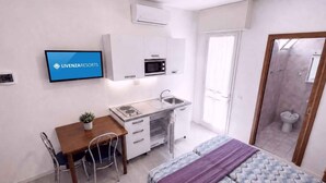 Ett kompakt hotellrum med ett litet pentry, en säng med mönstrat sänglinne, ett litet matbord och en tv som visar hotellets namn.