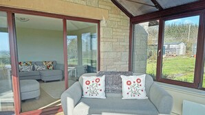 Ein modernes Wohnzimmer mit grauem Sofa, weißem Couchtisch und großen Fenstern mit Blick auf einen Garten und ein Steinhaus.