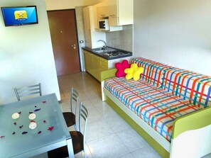 Un espacio habitable compacto con kitchenette, un sofá con cojines de colores, una mesa de comedor y un televisor.