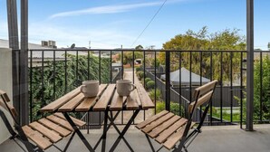 Un balcon avec une table et des chaises en bois, une rambarde en métal et une vue sur un jardin et des bâtiments.