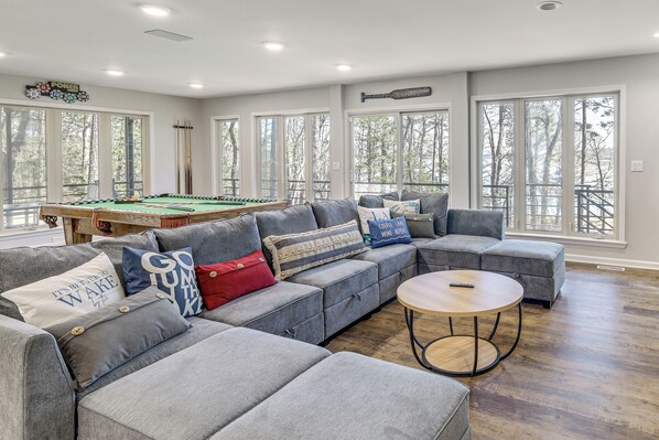 A modern living room with a pool table, grey sectional sofa, and large windows. - Hot Tub & Games Galore! Greers Ferry Lake Home (Fairfield Bay)