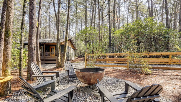 Ein rustikales Holzhäuschen mit Veranda, umgeben von einem Wald. Im Vordergrund stehen Adirondack-Stühle und eine Feuerstelle.