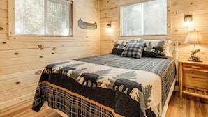 Ein Schlafzimmer einer Holzhütte mit einem Bett, zwei Fenstern, einem Nachttisch und einer Lampe.
