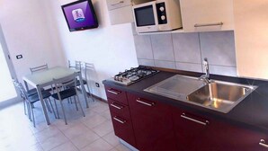 Eine kleine Küche mit Gasherd, Mikrowelle und Spüle aus Edelstahl. Es gibt einen Esstisch mit Stühlen und einen an der Wand befestigten Fernseher.